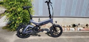 16インチのスタイリッシュな車輪を備えた折りたたみ電動自転車「FUNVITA」は、緑に囲まれたコンクリートの道の金属フェンスの横に駐車され、快適な走行を約束します。