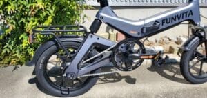 緑豊かな壁の近くの日当たりの良い歩道に駐車された、チェーンドライブと後部荷物ラックを備えたグレーの Funvita 16 インチ折り畳み電動自転車の拡大図。この多彩な乗り心地で快適な50キロ走行体験をお楽しみください。