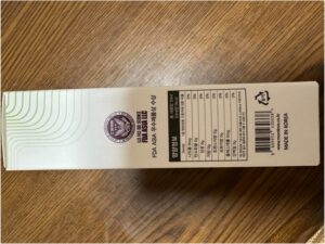 ベージュ色の商品箱に韓国語の文字と栄養成分表、そして英語で「MADE IN KOREA」と書かれています。ペットヘルスケア認証シールは、ペットの健康に焦点を当てていることを強調しています。箱は木の表面の上に置かれています。.