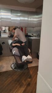 モダンで明るいサロンの洗面台で、ヘナトリートメントと洗髪をしている間、サロンの椅子にリクライニングしている人。背景にはキャビネット、鏡、サロン設備が見える。.