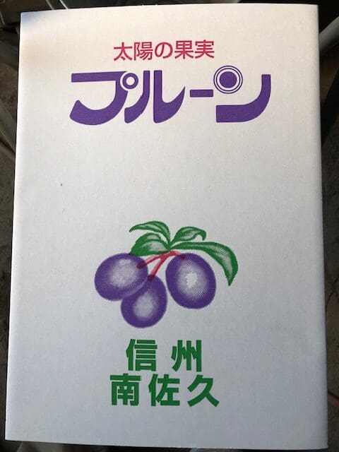日本語の文字が入った白いパッケージに、緑の葉をつけた3つの紫色の梅（プルーン）のイラストが描かれている。テキストは紫と緑が中心で、一部赤がアクセントとなっており、全体的にシンプルでクリーンなデザインとなっている。.
