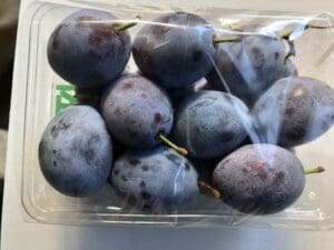 透明なプラスチックの容器に、茎が短く、白っぽい花をつけたサンプルーンプラムがいくつか入っている。プルーンプラムは白い表面に飾られ、予約販売に最適。.