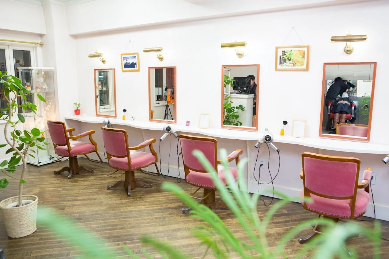 明るく整頓されたヘアサロンで、ショートカットのサービスを提供している。白い壁に鏡に向かってピンクの椅子が4脚。ドライヤーは各ステーションに設置され、緑の観葉植物が空間を飾り、2人の人物が映り込んでいる。.