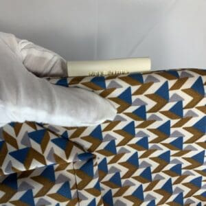 手袋をはめた手が、青、茶、白の幾何学模様の布地を持っている。生地の上に「MADE IN ITALY」と書かれた白いタグが部分的に見え、これがROBERTA PIERI TATAMI スモールトート イタリア製であることを示唆している。.
