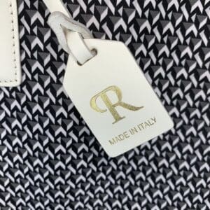 白と黒の幾何学模様の背景に、ゴールドの「R」の文字と「MADE IN ITALY」（イタリア製）の文字があしらわれた白いロベルタ・ピエリのラゲージタグが、ミニダッフにエレガンスを添えています。.