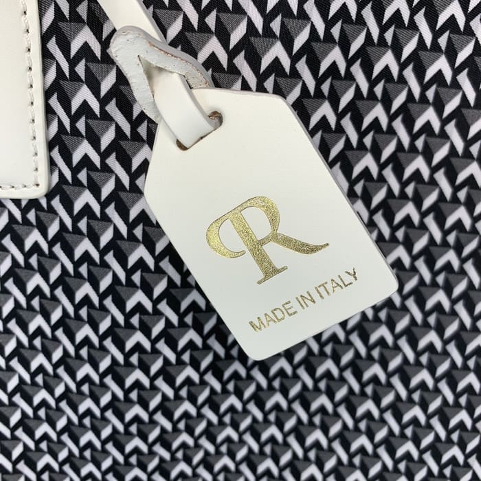 白と黒の幾何学模様の背景に、ゴールドの「R」の文字と「MADE IN ITALY」（イタリア製）の文字があしらわれた白いロベルタ・ピエリのラゲージタグが、ミニダッフにエレガンスを添えています。.