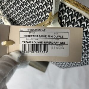 手袋をはめた手がベージュのROBERTA PIERIの商品タグを持ち、スタイル "ROBERTINA 2(DUE) MINI DUFFLE"、カラー "Tatami Lounge Supergray_OSS "など、ミニダッフルバッグのアイテム詳細を誇らしげにイタリア製で表示している。.
