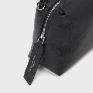シルバーのファスナー、「GIANNI CHIARINI FIRENZE」と白抜きで書かれた黒のレザータグが付いたGIANNI CHIARINI ALIFA ブラックテクスチャードレザーバッグのクローズアップを、明るい背景で撮影。.