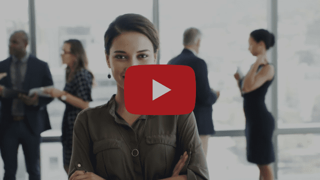 腕を組んで自信たっぷりに立ち、カメラに向かって微笑み、力強いパフォーマンスを体現する女性。ぼやけた背景には、大きな窓のそばで会話を交わす数人の人々。赤い再生ボタンのアイコンが画像に重なっている。.