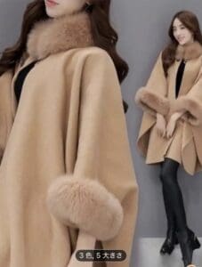 襟と袖口にファーのトリミングが施されたベージュレディースコートを着用し、黒いドレスと黒いタイツを合わせた女性。グレーの背景に立っている。.