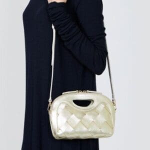 無地の淡い色を背景に、トップハンドルと白いショルダーストラップが付いたライトゴールドの編み目が特徴の「PELICO ビッグメッシュハンドバッグ スペイン製」を手にする黒い長袖ドレスの人物。.