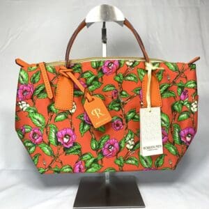 ピンクとパープルの花柄、グリーンの葉、ブラウンのハンドル、ゴールドの金具、ファスナー開閉式、タグが2つ付いた鮮やかなオレンジ色のロベルタ・ピエリ ミニダッフル ハンドバッグが、白を背景にスタンドに飾られている。イタリア製。.