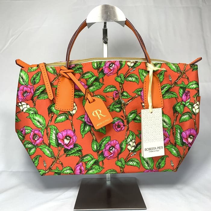ピンクとパープルの花柄、グリーンの葉、ブラウンのハンドル、ゴールドの金具、ファスナー開閉式、タグが2つ付いた鮮やかなオレンジ色のロベルタ・ピエリ ミニダッフル ハンドバッグが、白を背景にスタンドに飾られている。イタリア製。.