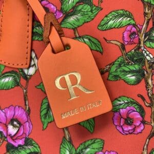 ミニダッフルバッグのピンクの花と緑の葉をモチーフにした赤い花柄の布地に取り付けられた、ゴールドのRとイタリア製テキストが入ったオレンジ色の革製タグ「ROBERTA PIERI」のクローズアップ。.
