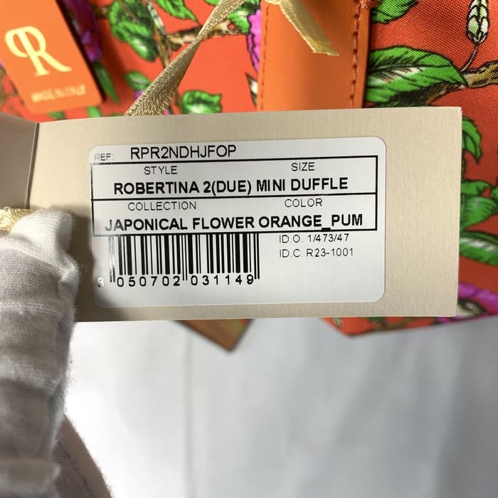 手袋をはめた手で持つ、スタイルROBERTINA 2(DUE) MINI DUFFLE in Japonical Flower Orange_Pumを示す商品タグのアップ。背景にROBERTA PIERIのカラフルな花柄のミニダッフル2WAYバッグが部分的に見える。.