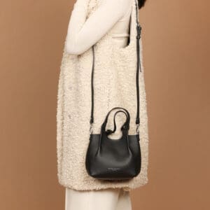 白いトップスとパンツの上に、クリーム色の質感のあるノースリーブのコートを着た人物が、茶色を背景に短いハンドルと長いショルダーストラップが付いた黒のレザーハンドバッグ「ジャンニ・キアリーニ ドゥア XS」を手にしている。.