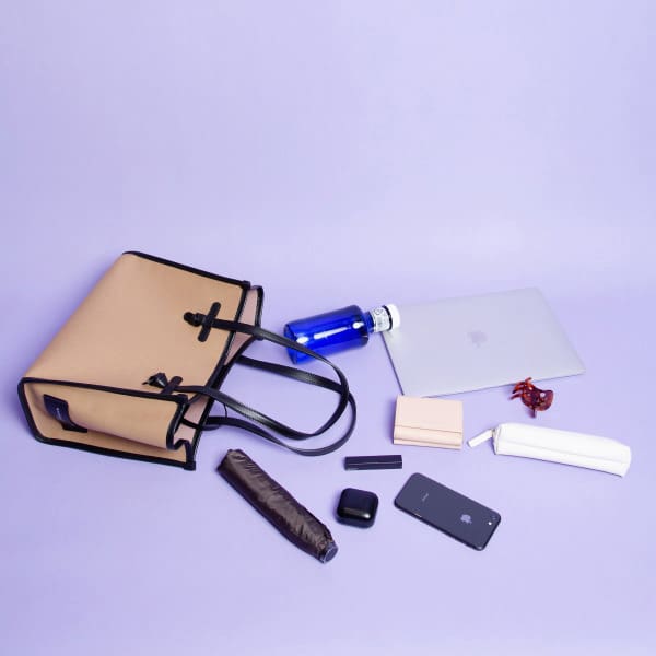 ベージュのGIANNI CHIARINIのトートバッグ「イタリア製」はラベンダー色の表面に置かれ、中身は青い水筒、ノートパソコン、サングラス、財布、筆箱、傘、リップスティック、AirPods、そして黒いスマートフォン。.