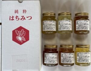 日本語の文字と赤い蜂のアイコンが描かれた箱の隣には、金色の蓋と日本語のラベルが貼られた八ヶ岳高原の小さな瓶が6つ、白い表面に詰め合わせのように美しく並べられている。.
