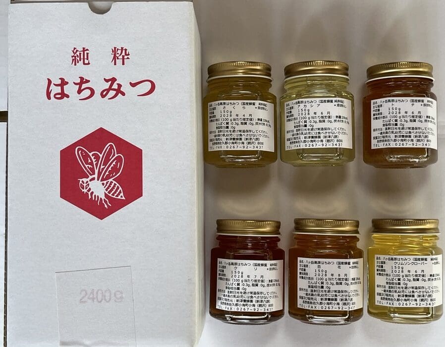 日本語の文字と赤い蜂のアイコンが描かれた箱の隣には、金色の蓋と日本語のラベルが貼られた八ヶ岳高原の小さな瓶が6つ、白い表面に詰め合わせのように美しく並べられている。.