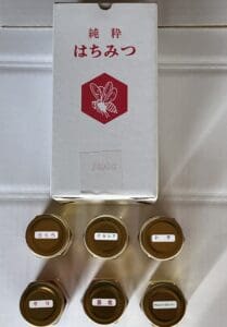 赤い蜂のイラストが描かれた日本語のラベルが貼られた箱が、八ヶ岳高原はちみつの5つの小瓶の上に置かれている。それぞれの瓶には日本語のラベルが貼られ、ユニークな風味の蜂蜜の詰め合わせが紹介されている。.
