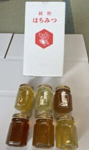 黄色と琥珀色の色合いが異なる6つの「ちみつ」の瓶が、「2400g」「八ヶ岳高原詰め合わせ」と赤い蜂のマークが入った日本語のラベルが貼られた白い箱の前に2列に並べられている。.