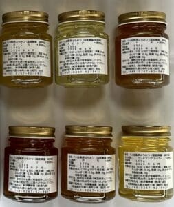 八ヶ岳高原はちみつの6つの小さな瓶が2列に並べられ、それぞれに金色の蓋と日本語の文字が書かれた白いラベルが付いている。瓶入り詰め合わせは、淡い琥珀色から濃い琥珀色までの蜂蜜の色合いを見せている。.