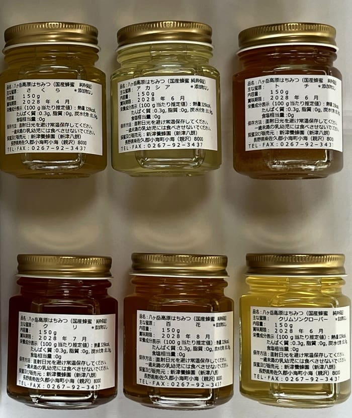 八ヶ岳高原はちみつの6つの小さな瓶が2列に並べられ、それぞれに金色の蓋と日本語の文字が書かれた白いラベルが付いている。瓶入り詰め合わせは、淡い琥珀色から濃い琥珀色までの蜂蜜の色合いを見せている。.