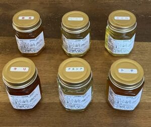 金色の蓋が付いた八ヶ岳高原はちみつの瓶が6つ、木製の表面に並べられている。それぞれの瓶には日本語の白いラベルと色のついた蓋のラベルが貼られ、蜂蜜好きにはたまらない詰め合わせセットです。.