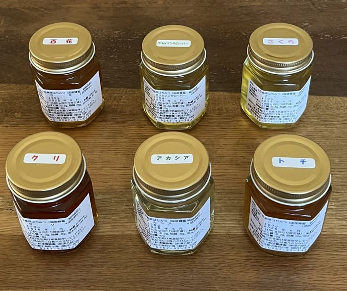 金色の蓋が付いた八ヶ岳高原はちみつの瓶が6つ、木製の表面に並べられている。それぞれの瓶には日本語の白いラベルと色のついた蓋のラベルが貼られ、蜂蜜好きにはたまらない詰め合わせセットです。.