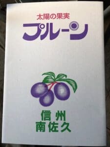 日本語の文字と、緑の葉をつけた3つの紫色のプルーンのイラスト、さらに紫、赤、緑の太字の文字が施された白いパッケージ。 「太陽の果実プルーン サマーキュート（中玉種）」のデザインは、その季節ならではの魅力を強調しており、8月17日頃から2週間にわたって予定されている出荷期間に先立ち、DS予約販売に最適だ。.
