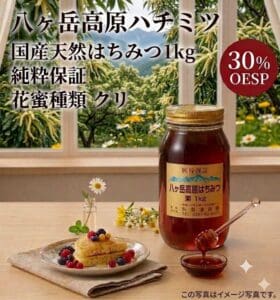 木のテーブルの上には、ベリーをトッピングしたパンケーキの皿の横に、スプーンが添えられた「八ヶ岳高原 国産天然はちみつ 1kg 純粋保証 花蜜の種類：クリ」の瓶が置かれている。画像には、日本語の文字と「OESP 30%」と記された赤いラベルが見える。.