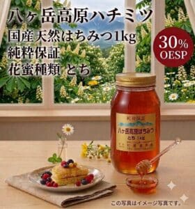 「八ヶ岳高原 国産天然はちみつ 1kg 純粋保証（花蜜の種類：トチ）」の瓶とハチミツ用スプーンが、フルーツをトッピングしたパンケーキの乗ったお皿の隣のテーブルに置かれている。背景には花や緑が飾られており、画像には日本語の文字と赤い「30% OESP」のラベルが見える。.