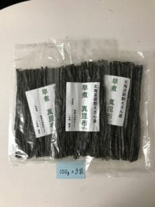 白い台の上に、「北海道恵山」や「早煮昆布」といった日本語の表記がされた透明なプラスチック袋3袋（北海道恵山 早煮昆布、100g入り×3袋セット）が並べられています。 その下にある青いメモには「100g×3袋」と書かれており、このプレミアム昆布セットの各袋には100グラムが入っていることを示しています。.