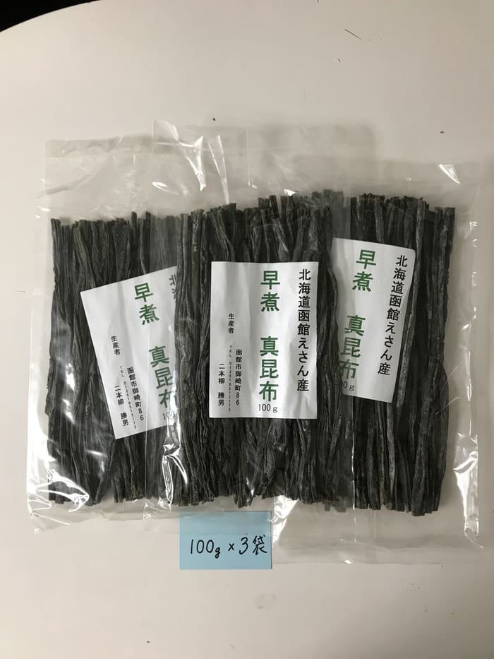 白い台の上に、「北海道恵山」や「早煮昆布」といった日本語の表記がされた透明なプラスチック袋3袋（北海道恵山 早煮昆布、100g入り×3袋セット）が並べられています。 その下にある青いメモには「100g×3袋」と書かれており、このプレミアム昆布セットの各袋には100グラムが入っていることを示しています。.