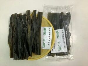 黄色いトレイの上に、乾燥昆布（具体的には「北海道恵山 真昆布（だし用）」）が数枚並べられており、その横には同じ商品の透明な袋が2つ置かれています。それぞれの袋には「100g」と表示され、日本語のラベルが貼られています。このセットには、100g入りの袋が2袋含まれています。.