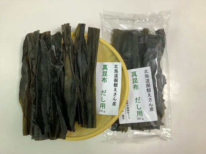 黄色いトレイの上に、乾燥昆布（具体的には「北海道恵山 真昆布（だし用）」）が数枚並べられており、その横には同じ商品の透明な袋が2つ置かれています。それぞれの袋には「100g」と表示され、日本語のラベルが貼られています。このセットには、100g入りの袋が2袋含まれています。.