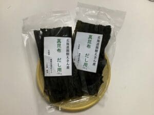 白い背景を背に、小さな丸い竹のトレイの上に、北海道産エサンマコンブ（出汁用）が2パック（各100g）置かれている。.