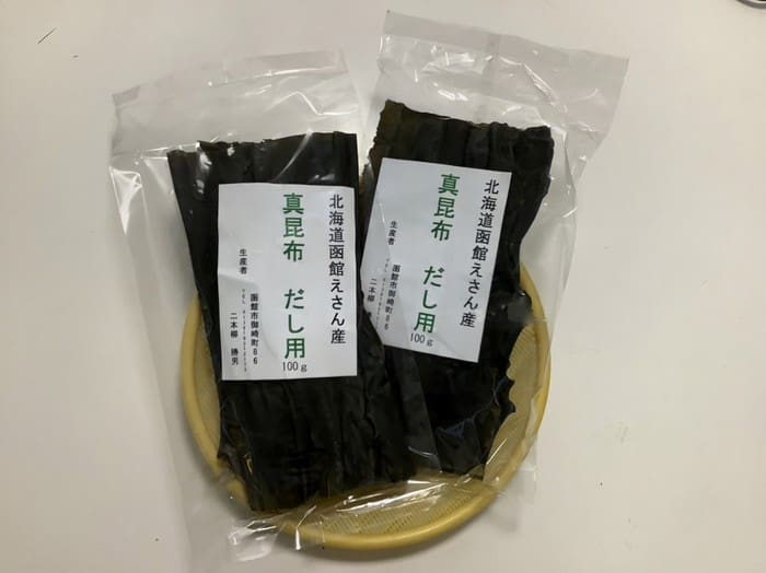 白い背景を背に、小さな丸い竹のトレイの上に、北海道産エサンマコンブ（出汁用）が2パック（各100g）置かれている。.