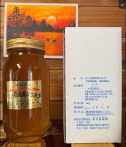 金色のラベルが貼られた「八ヶ岳高原 国産天然はちみつ 1kg 純粋保証」の瓶が、花の源が「とち」であると日本語で記された白い箱の隣に置かれている。背景には、木々や鹿が描かれた夕日の絵があり、この純粋な国産はちみつが採れる地域の自然の美しさを映し出している。.